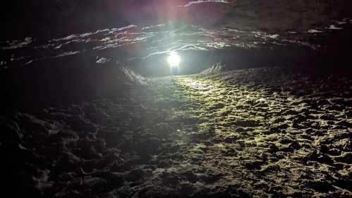 A dimly lit cave with a bright light at the end, illuminating the sandy ground and rocky walls.