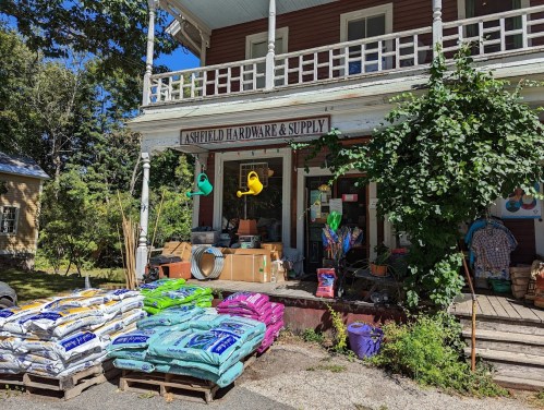 A quaint hardware store with colorful bags of supplies outside, surrounded by greenery and a welcoming porch.