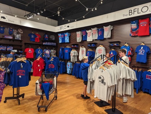 A retail store displaying various sports apparel in blue and red, with customers browsing shirts and merchandise.