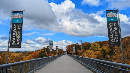 A wide walkway with banners reading "Walkway Over the Hudson," surrounded by colorful autumn trees and a blue sky.