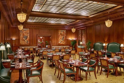 Elegant restaurant interior with wooden paneling, green leather chairs, and stained glass ceiling, set for dining.