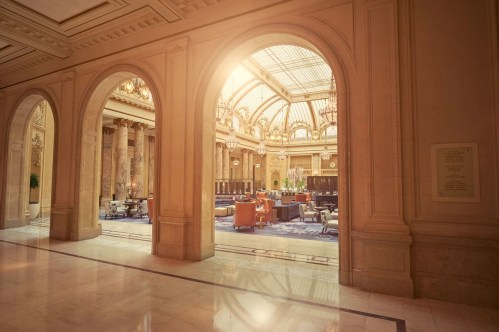 Elegant interior view of a grand hall with arches, sunlight streaming through a glass ceiling, and plush seating.