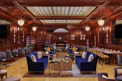 Elegant lounge with dark wood paneling, plush blue sofas, and a grand piano, featuring soft lighting and stylish decor.