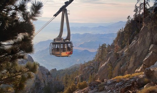 A cable car glides over a mountainous landscape, surrounded by trees and distant peaks under a soft sunset sky.