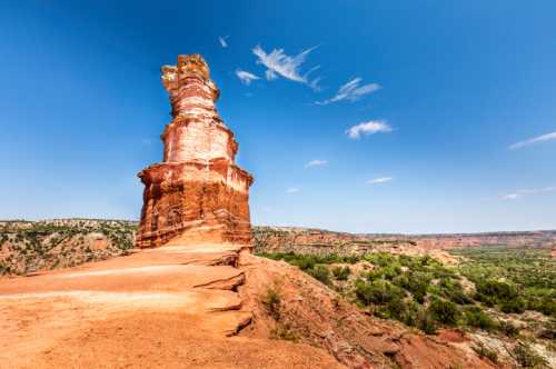 A tall, reddish rock formation stands against a clear blue sky, surrounded by green hills and rugged terrain.