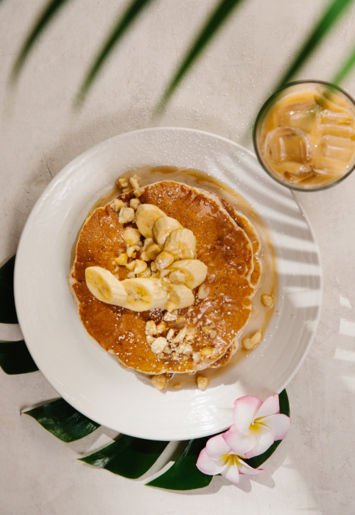 A plate of pancakes topped with banana slices and nuts, drizzled with syrup, beside a glass of iced coffee.