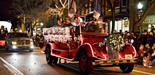 A festive red fire truck adorned with lights and decorations, parading down a street with onlookers enjoying the event.