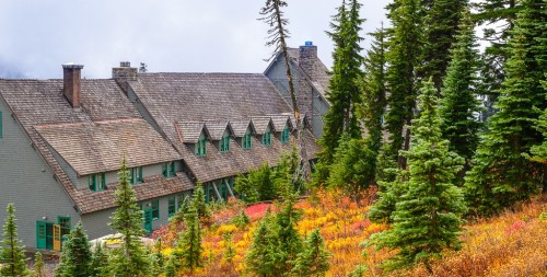 A rustic lodge nestled among evergreen trees and colorful autumn foliage under a cloudy sky.