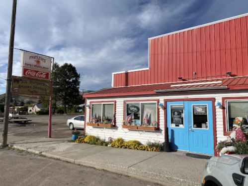 A small, red-roofed diner with a blue door, surrounded by flowers and a sign for burgers and steak.
