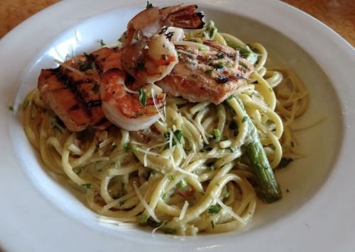 A plate of pasta topped with grilled shrimp, chicken, asparagus, and sprinkled with cheese and herbs.
