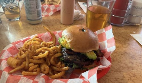A juicy burger with lettuce and pickles, served with curly fries and a glass of beer on a checkered table.
