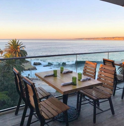 A seaside dining area with a wooden table and chairs, overlooking the ocean at sunset. Palm trees in the background.