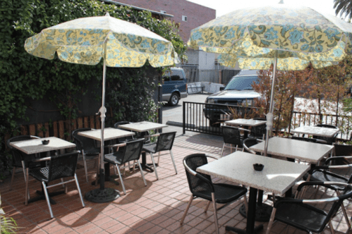 Outdoor dining area with tables and colorful umbrellas, surrounded by greenery and a parked car in the background.