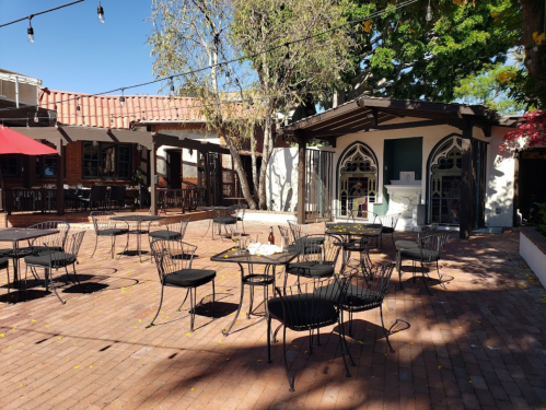 Outdoor patio with empty tables and chairs, surrounded by trees and buildings, under string lights.