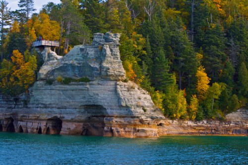A rocky cliff with vibrant autumn foliage and a wooden lookout tower above a calm, turquoise lake.