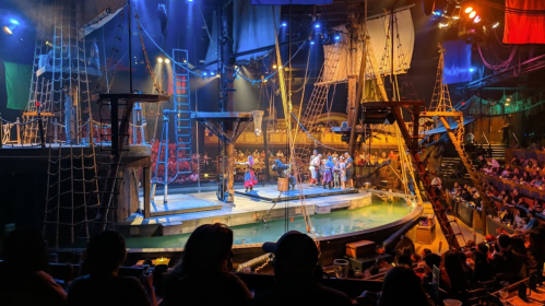 A lively pirate-themed stage with actors performing on a ship, surrounded by an audience in a dimly lit venue.