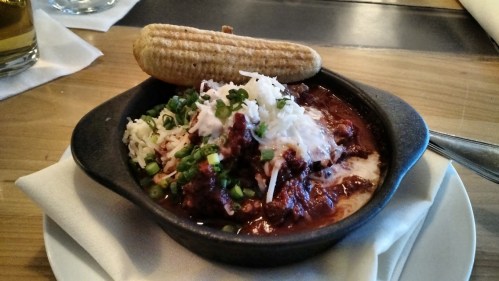 A small black bowl filled with chili topped with cheese and green onions, accompanied by a corn muffin on the side.
