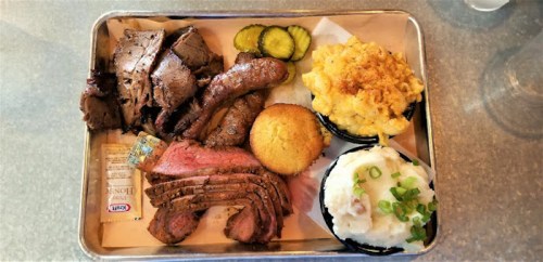A metal tray filled with sliced meats, pickles, cornbread, macaroni and cheese, and mashed potatoes topped with green onions.