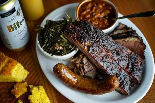A plate of barbecue featuring brisket, sausage, collard greens, baked beans, and cornbread, with a beer on the side.