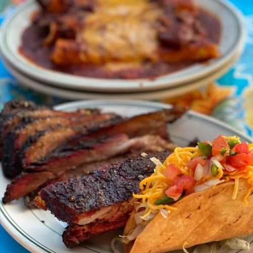 A plate featuring grilled ribs, a taco topped with cheese and pico de gallo, with enchiladas in the background.
