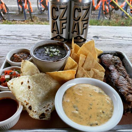 A tray of food featuring chips, salsa, black beans, a beef steak, and two cans of Czech-style pilsner beer.