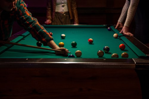 A person plays pool, aiming at colorful balls on a green felt table in a dimly lit setting.