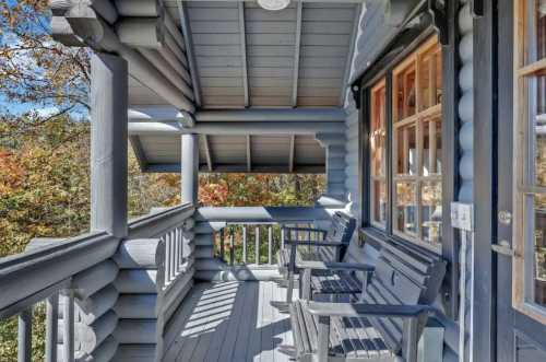 A log cabin porch with wooden chairs, surrounded by trees and autumn foliage under a clear blue sky.