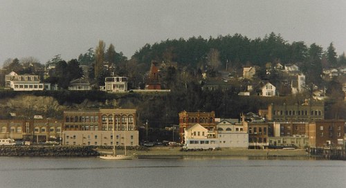 A scenic view of a waterfront town with historic buildings and a hillside covered in trees.
