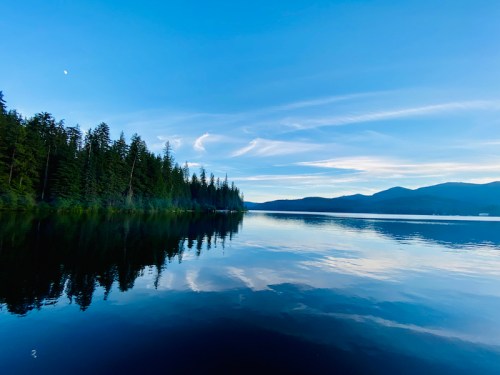 A serene lake surrounded by trees, reflecting a clear blue sky and distant mountains.