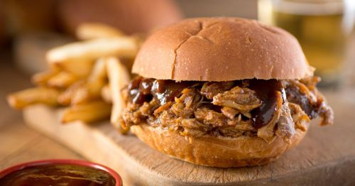close up of pulled pork barbecue sandwich next to a pile of fries on a wooden cutting board