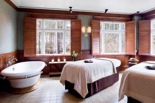 A serene spa room with two massage tables, a freestanding bathtub, and large windows with wooden shutters.
