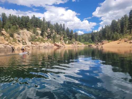 A serene lake surrounded by trees and rocky cliffs, with a person kayaking on the calm water under a blue sky.