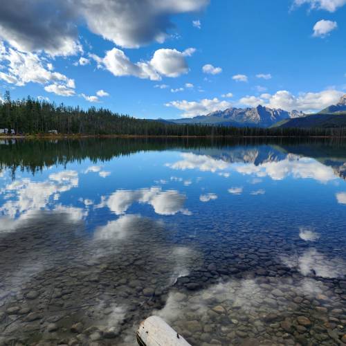 A serene lake reflecting clouds and mountains, surrounded by lush trees under a bright blue sky.