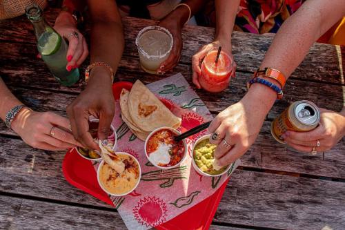 A group of hands reaching for colorful Mexican dishes on a wooden table, with drinks in the background.