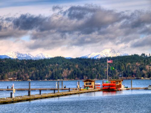 A scenic view of a lake with a dock, colorful kayaks, and mountains in the background under a cloudy sky.