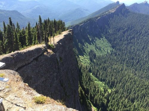 A rocky cliff overlooks a lush green valley, surrounded by dense forests and distant mountains under a clear blue sky.
