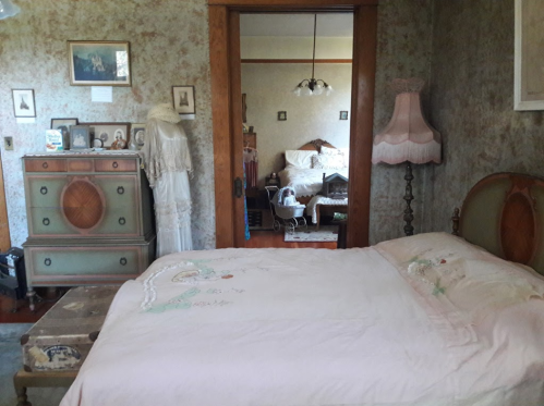 A vintage bedroom with a pink bedspread, antique furniture, and soft lighting, featuring a doorway to another room.