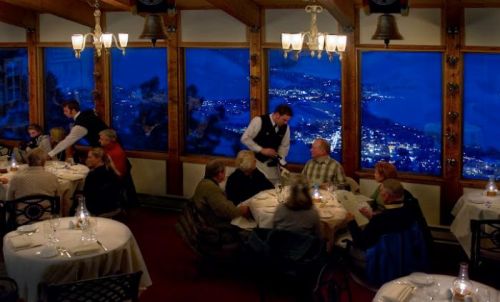 A cozy restaurant with diners enjoying a meal, featuring large windows showcasing a night view of city lights.