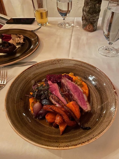 A plate of sliced duck breast on a bed of orange puree, garnished with roasted vegetables and a berry sauce.