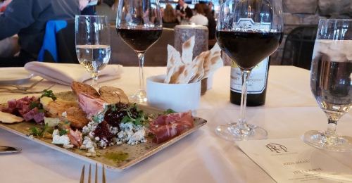 A gourmet platter with meats, cheeses, and crackers, accompanied by glasses of red wine and water on a dining table.