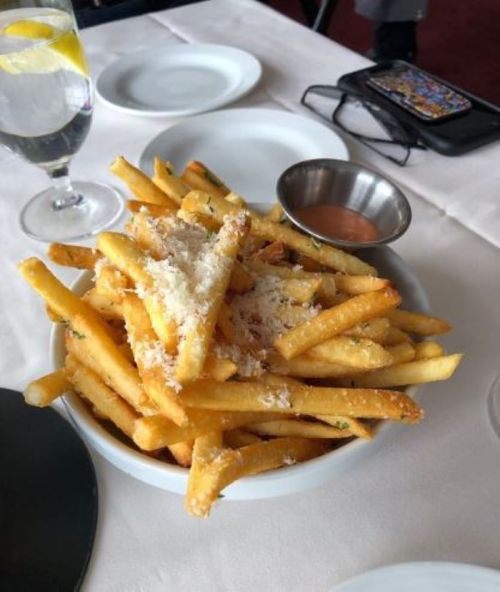 A bowl of golden fries topped with grated cheese, served with a small bowl of dipping sauce and a glass of water.