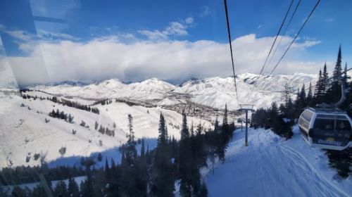 A scenic view from a gondola, overlooking snow-covered mountains and a valley under a blue sky with clouds.