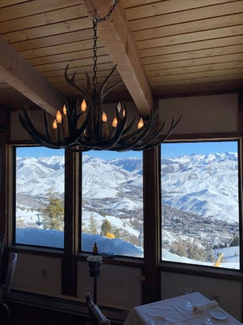 A rustic chandelier hangs in a cozy restaurant with large windows showcasing snowy mountain views.