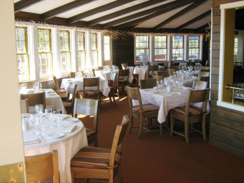 A cozy restaurant interior with tables set for dining, featuring white tablecloths and large windows.