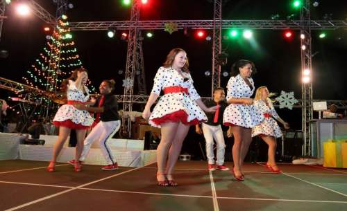 A group of dancers in polka dot outfits perform on stage with festive lights and a Christmas tree in the background.