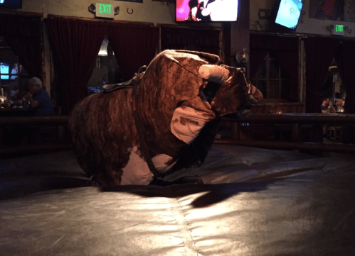 A mechanical bull sits on a padded surface in a dimly lit bar, with people visible in the background.