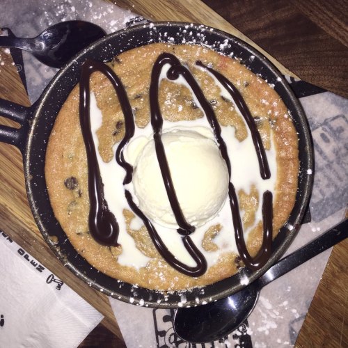 A warm chocolate chip cookie in a skillet, topped with vanilla ice cream, chocolate drizzle, and powdered sugar.