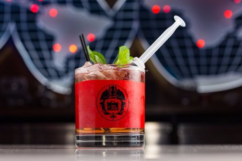 A cocktail in a glass with a red label, garnished with mint and served with a syringe for a unique presentation.