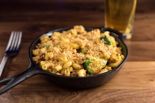 A cast-iron skillet filled with macaroni and cheese topped with breadcrumbs and broccoli, alongside a glass of beer.