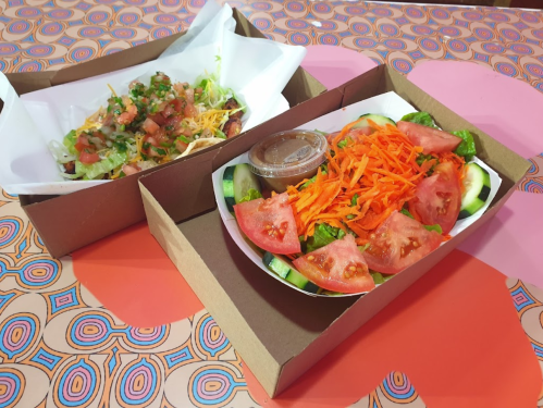 Two takeout boxes: one with a salad topped with carrots and tomatoes, the other with nachos and fresh toppings.